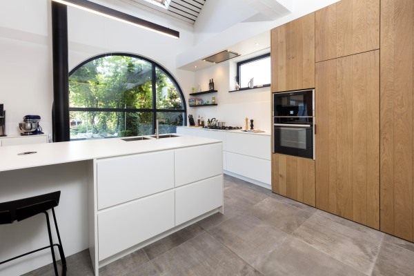 Strakke keuken met wit keukeneiland met spoelgedeelte en kastenwand met gasvuur en kolomkasten eik gebogen raam